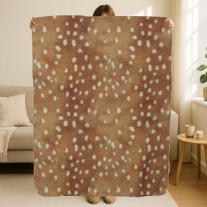 May include: A brown and white blanket with a pattern resembling deer fur. The blanket is held up, showcasing its texture and design. The blanket's color palette includes shades of brown and white.