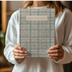 以下が含まれることがあります： 緑、ラベンダー、ピンクの色合いのチェック柄のハードカバーノート。ノートの表紙には、薄いベージュ色で「Gratitude 2026」の文字が書かれています。チェック柄には、花や葉のような要素が含まれています。