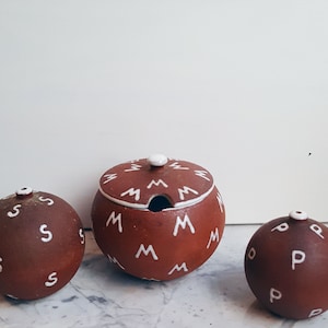 May include: Three ceramic salt and pepper shakers with lids. The shakers are brown with white letters on them. The letters on the shakers are "S", "M", and "P".