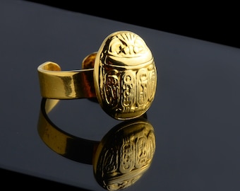 May include: A gold ring with a scarab beetle design. The beetle is detailed with hieroglyphics and has a raised, textured surface.