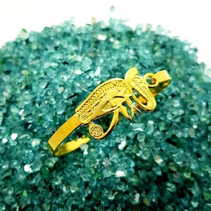 May include: A gold-colored bracelet featuring an Egyptian Eye of Horus design. The bracelet is set against a backdrop of small, teal-colored stones. The bracelet's design includes intricate details and a curved shape.
