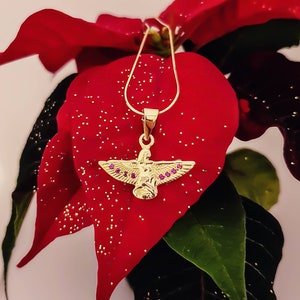 May include: A gold pendant necklace featuring a winged angel design. The angel has purple gemstones on its wings and body. The necklace is displayed on a red poinsettia.