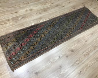 May include: A long, rectangular rug with a geometric diamond pattern in shades of red, blue, green, and gold. The rug has a dark brown border and fringe on the ends. The rug is placed on a light-colored wooden floor.