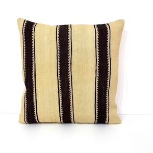 May include: A square throw cushion featuring a woven texture with alternating vertical stripes. The design consists of wide, light beige panels separated by narrower, dark brown stripes that have a distinct, repeating zigzag edge pattern.