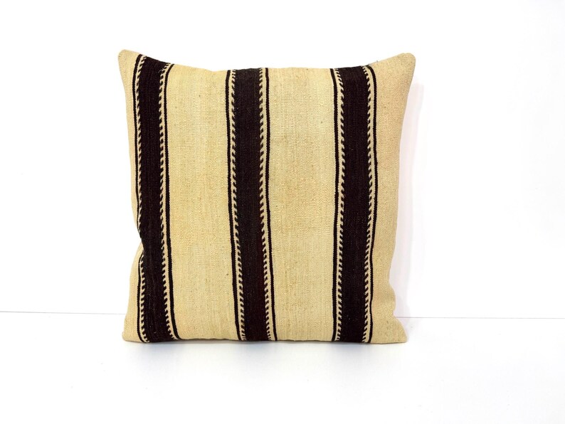 May include: A square throw cushion featuring a woven texture with alternating vertical stripes. The design consists of wide, light beige panels separated by narrower, dark brown stripes that have a distinct, repeating zigzag edge pattern.