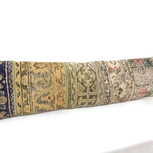 May include: Long, rectangular cushion with a patchwork design. The cushion features a variety of colours, including navy blue, gold, green, and pink. The design appears to be made from sections of vintage rugs, creating a unique and textured look.