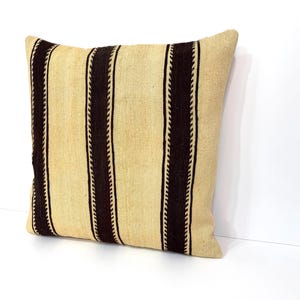 May include: A square throw cushion featuring a textured beige base with three vertical dark brown stripes. Each dark brown stripe is bordered by a thin, lighter-coloured zigzag or chevron pattern, creating a distinct contrast against the neutral fabric.