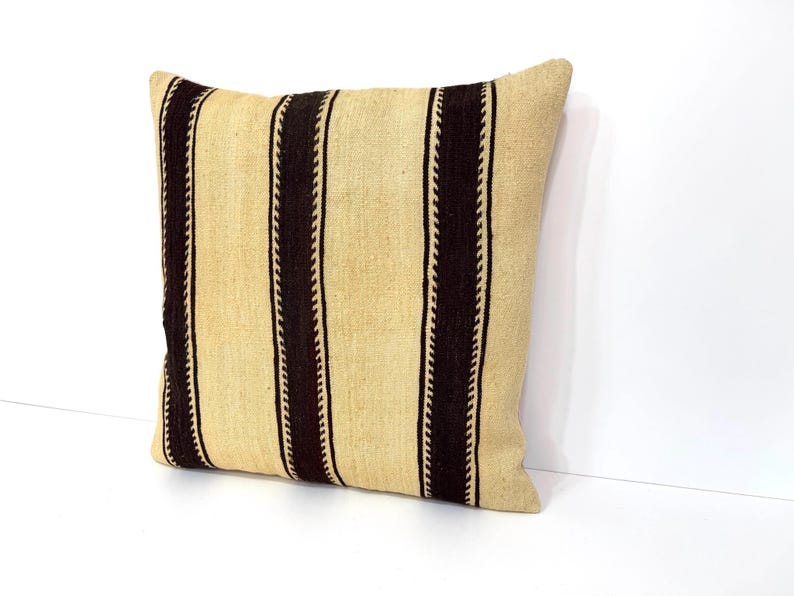 May include: A square throw cushion featuring a textured beige base with three vertical dark brown stripes. Each dark brown stripe is bordered by a thin, lighter-coloured zigzag or chevron pattern, creating a distinct contrast against the neutral fabric.