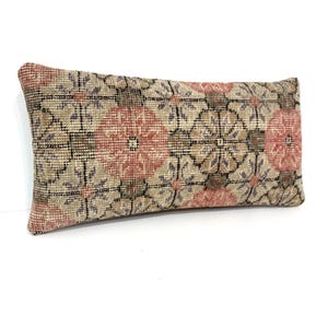 Puede incluir: Un cojín lumbar rectangular con una funda de tejido textil que presenta un patrón floral geométrico repetitivo en tonos apagados de rosa salmón, beige, marrón y gris oscuro. La tela tiene una apariencia texturizada con detalles de tejido.