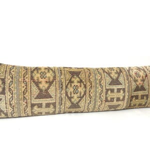 May include: A long, rectangular pillow with a woven, patterned design. The pillow features a mix of brown, beige, and green hues with geometric and floral motifs. The pillow is likely used for decorative purposes.