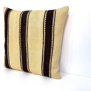 May include: A square throw cushion featuring a textured, light beige fabric base with three prominent vertical dark brown stripes. Each dark brown stripe is bordered by a thin, zigzag-patterned line, creating a distinct contrast against the neutral background.