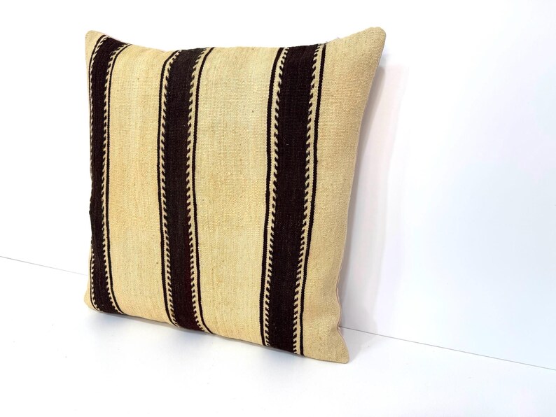 May include: A square throw cushion featuring a textured, light beige fabric base with three prominent vertical dark brown stripes. Each dark brown stripe is bordered by a thin, zigzag-patterned line, creating a distinct contrast against the neutral background.