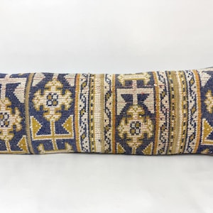 May include: A rectangular lumbar cushion featuring a woven textile pattern in shades of navy blue, mustard yellow, and cream. The design consists of repeating geometric motifs and vertical striped bands across the entire surface of the fabric.