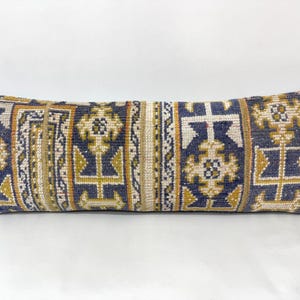 Puede incluir: Un cojín lumbar rectangular con una funda de tejido con un patrón geométrico repetitivo en tonos azul marino, amarillo mostaza y crema. El diseño consta de paneles verticales con motivos florales y de cruz estilizados.