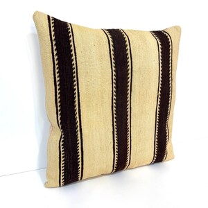 May include: A square throw cushion featuring a woven texture with a pattern of wide, dark brown vertical stripes alternating with wider beige sections. The dark brown stripes are bordered by thin, intricate, lighter-coloured zigzag lines.