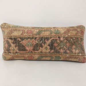 May include: A rectangular cushion with a distressed, vintage appearance. It has a central band of dark brown with floral patterns in red, green, and beige. The edges and background are a lighter beige, creating a layered effect. A decorative accent for a sofa or bed.