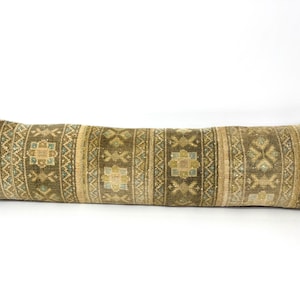 May include: A long, rectangular cushion with a woven, patterned design. The colour palette includes olive green, beige, and blue accents. The pattern features geometric shapes and floral motifs, creating a vintage or bohemian aesthetic.