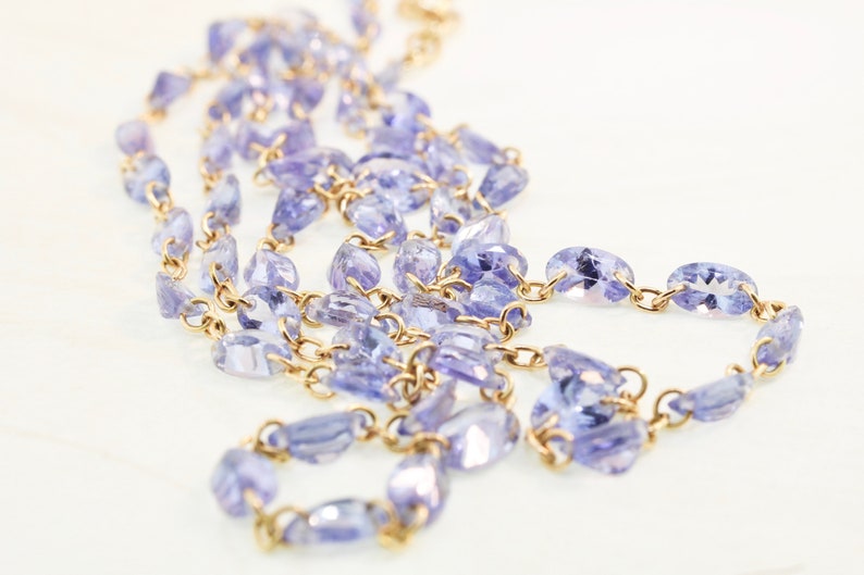May include: A gold chain necklace with purple gemstone beads. The chain is made of small, linked metal pieces. The beads are oval-shaped and have a slightly irregular shape.