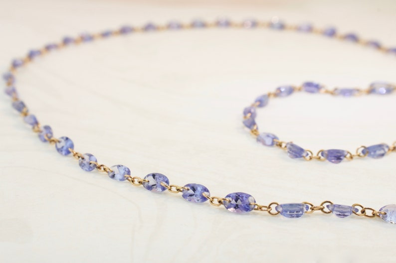 May include: A delicate gold chain necklace with oval-shaped amethyst gemstones. The necklace is laid out on a white surface.