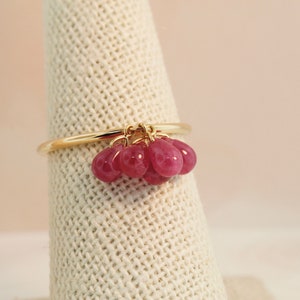 May include: A gold ring featuring a cluster of small, teardrop-shaped, bright pink gemstones. The ring is displayed on a textured, off-white surface, with a soft, neutral background.