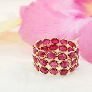 May include: A gold ring with multiple rows of oval-shaped pink gemstones. The ring is set against a background of pink flower petals.