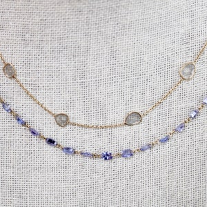 May include: Two gold necklaces with gemstones. The top necklace has four clear, faceted stones and a gold chain. The bottom necklace has a chain of small, light blue stones.