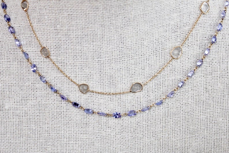 May include: Two gold necklaces with gemstones. The top necklace has four clear, faceted stones and a gold chain. The bottom necklace has a chain of small, light blue stones.