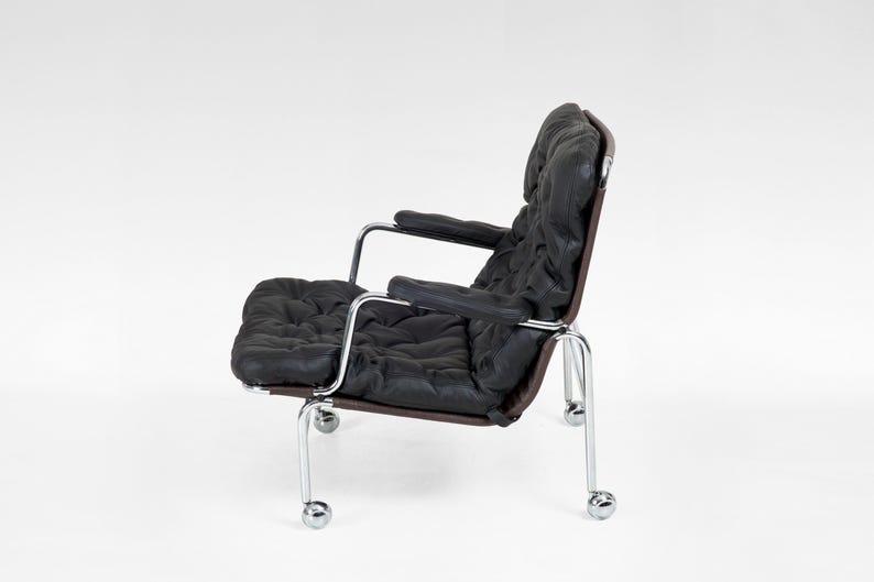 May include: Black leather armchair with a tufted seat and backrest. The chair has a dark brown frame and chrome arms and legs with rolling casters. The chair is set against a white background.