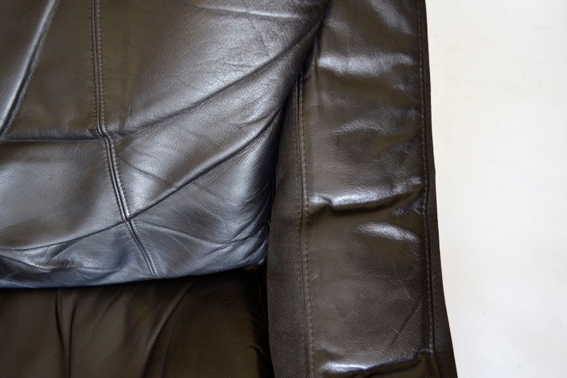 May include: Close-up of a section of a black leather sofa. The leather is stitched in a geometric pattern. The armrest is visible on the right side, with a seam running vertically. The background is white.