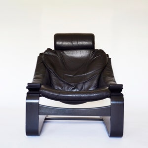 May include: Black leather armchair with a high back and headrest. The chair has a curved wooden frame and armrests. The design is modern and minimalist, with a focus on comfort and style. The chair is set against a white background.