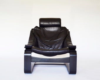 May include: Black leather armchair with a high back and headrest. The chair has a curved wooden frame and armrests. The design is modern and minimalist, with a focus on comfort and style. The chair is set against a white background.