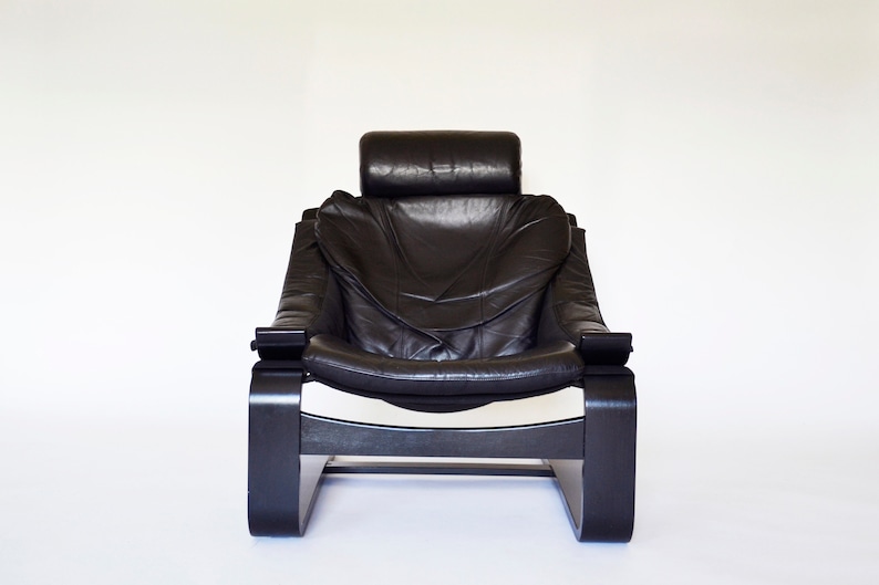 May include: Black leather armchair with a high back and headrest. The chair has a curved wooden frame and armrests. The design is modern and minimalist, with a focus on comfort and style. The chair is set against a white background.