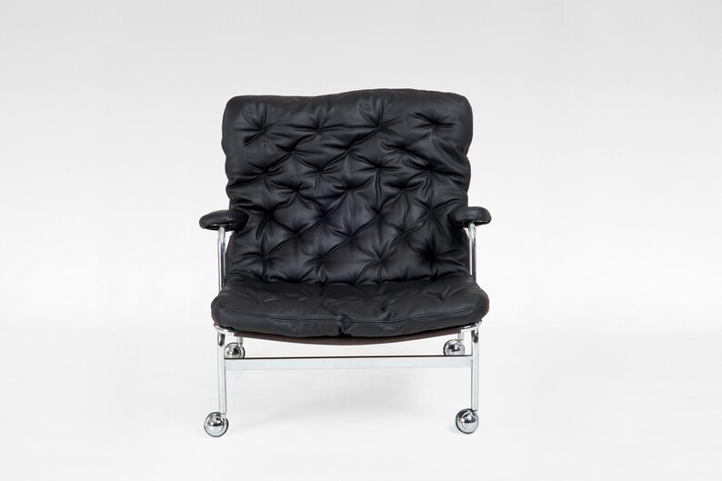 May include: Black leather armchair with a tufted back and seat. The chair has chrome arms and a chrome base with four small wheels. The chair is designed for comfort and style, suitable for a living room or office space.