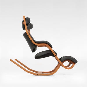 Puede incluir: Una silla mecedora de cuero negro con un marco de madera. La silla tiene un respaldo alto y un reposapiés. La silla está diseñada para una postura ergonómica.