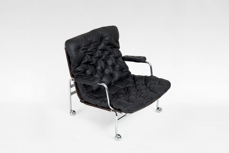 May include: Black leather armchair with a tufted back and seat. The chair has a chrome frame and armrests. The chair sits on four small chrome wheels. The chair is a modern design.