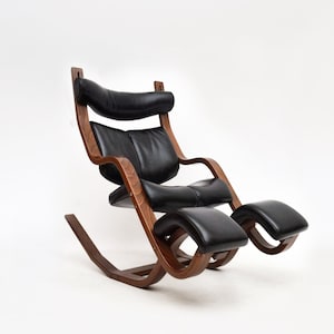 Puede incluir: Un sillón mecedora de cuero negro con reposapiés. La silla tiene un marco de madera curvado y un reposacabezas acolchado.