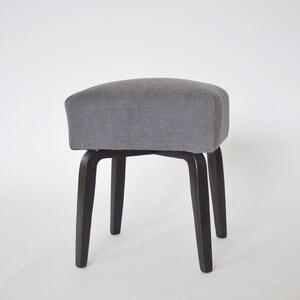 May include: A gray upholstered stool with four black wooden legs. The stool has a square seat and a simple design.