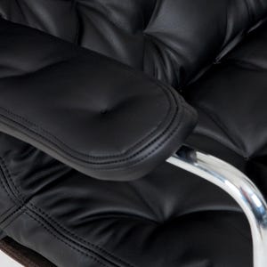 May include: Close-up of a black leather office chair with a tufted back and seat. The armrest is also black leather with a stitched edge. A chrome arm support is visible.
