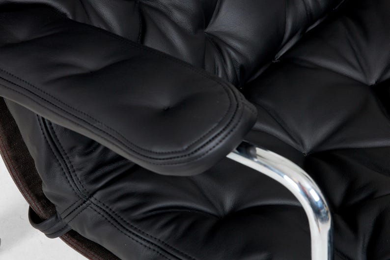 May include: Close-up of a black leather office chair with a tufted back and seat. The armrest is also black leather with a stitched edge. A chrome arm support is visible.