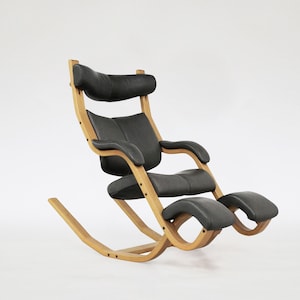 Puede incluir: Una silla mecedora de cuero negro con estructura de madera y reposapiés. La silla tiene un respaldo alto y brazos curvos.