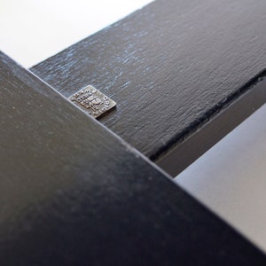 May include: Close-up of two intersecting black wooden beams. A small, square metal tag is affixed to one beam, bearing etched text. The beams have a textured surface and are set against a white background.