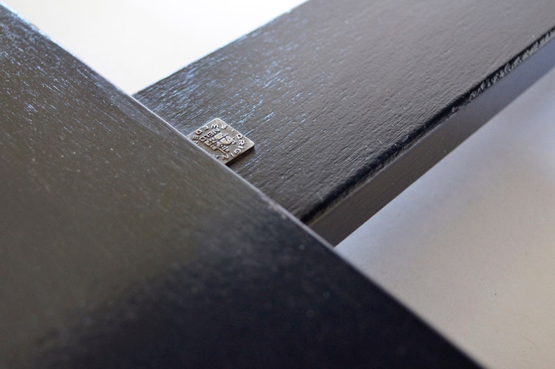 May include: Close-up of two intersecting black wooden beams. A small, square metal tag is affixed to one beam, bearing etched text. The beams have a textured surface and are set against a white background.