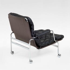 May include: Modern black leather armchair with a tufted seat and backrest, set against a brown fabric backing. The chair features a chrome frame and small caster wheels for mobility. A stylish piece of furniture.