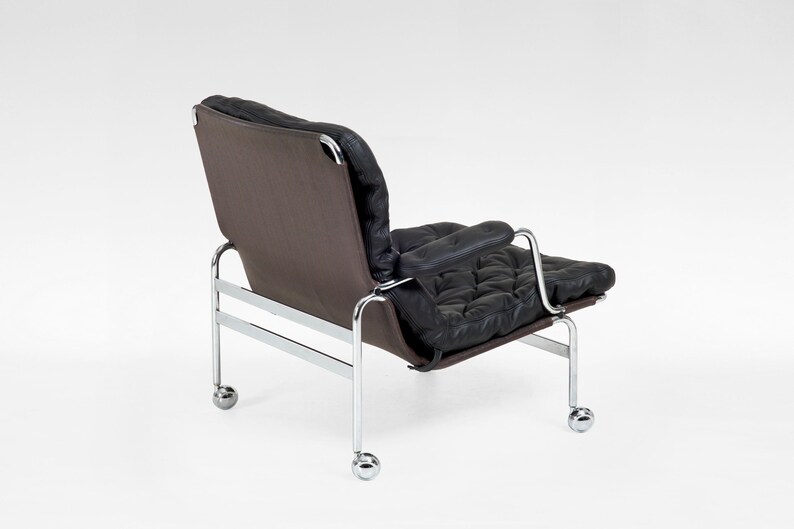 May include: Modern black leather armchair with a tufted seat and backrest, set against a brown fabric backing. The chair features a chrome frame and small caster wheels for mobility. A stylish piece of furniture.