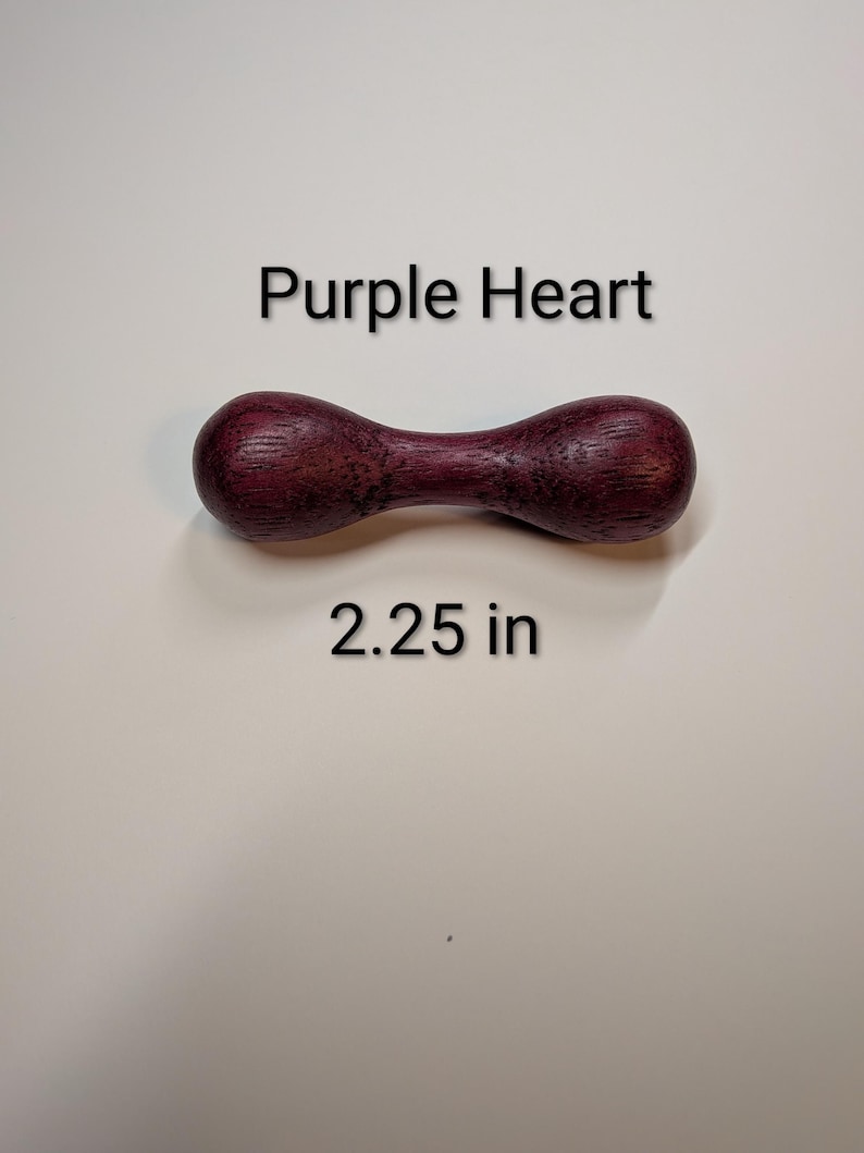 May include: A wooden, hourglass-shaped object with a deep purple hue. The text "Purple Heart" and "2.25 in" are displayed. The wood grain is visible, adding texture to the piece.