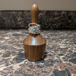 May include: A dark brown wooden ring holder with a tapered cylindrical shape. Three rings are stacked on the post: a thin silver band, a silver ring featuring a flower design with a small turquoise stone in the center, and a gold chain link ring.