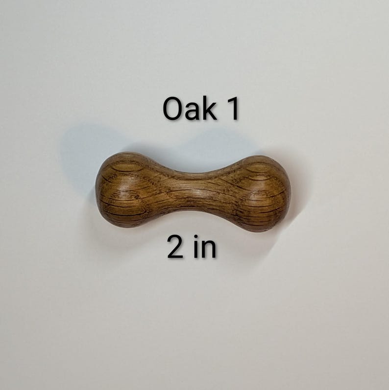May include: A wooden dumbbell-shaped object made of oak. The object is brown with visible wood grain and the text "Oak 1" and "2 in" are printed on the image.