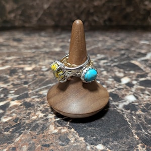 May include: A stack of three silver rings displayed on a smooth, conical wooden stand. One ring features a yellow and black patterned stone, another has a bright turquoise oval stone, and the third is a simple textured silver band.