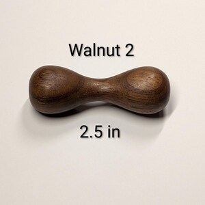 May include: A dark brown, wooden dumbbell-shaped object. The text "Walnut 2" is above the object, and "2.5 in" is below it. The object is approximately 2.5 inches long.