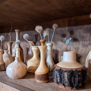 Puede incluir: Una colección de jarrones de madera hechos a mano de varias formas y tamaños, expuestos en una estantería de madera. Algunos jarrones presentan detalles de corteza natural, y varios contienen arreglos florales secos. Los tonos de madera varían.